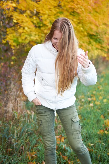 Tempo de outono. Retrato de jovem rindo menina positiva com cabelo comprido natural bonito vestindo jaqueta branca e calça verde. Árvores de outono amarelas sobre fundo.