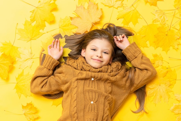 Tempo de outono Uma garotinha em um suéter de malha encontra-se em um fundo amarelo com folhas de bordo caídas
