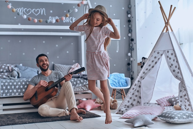 Tempo despreocupado juntos. Pai alegre tocando violão para sua filha e sorrindo enquanto passa o tempo livre em casa