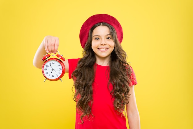 Tempo menina no chapéu estilo francês menina feliz com cabelo longo encaracolado na boina beleza cabeleireiro criança com despertador moda atemporal criança parisiense em amarelo bom dia preparação matinal