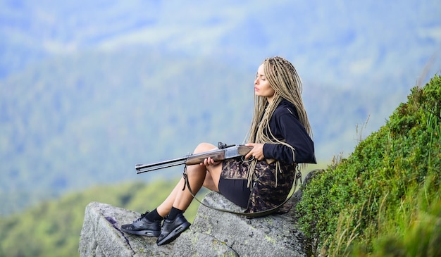 Temporada de caza Fondo de paisaje de montañas de cazador Guerrero sexy Concepto de puntería Chica amazona sentada en un acantilado preparar arma para cazar Mujer atractiva pelo largo cara bonita sostener rifle para cazar