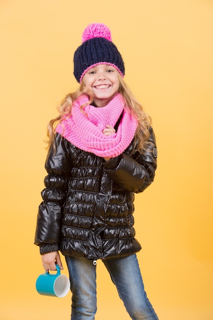 Temporada de otoño relajarse concepto. Niño con sonrisa de taza azul sobre fondo naranja. Bebida caliente en clima frío. Chica con sombrero, bufanda rosa, chaqueta negra con taza. Pausa para el té o el café.