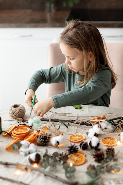 Temporada de vacaciones de invierno niño niña pequeña haciendo guirnaldas navideñas hechas a mano decoraciones para el hogar con materiales ecológicos naturales Vertical