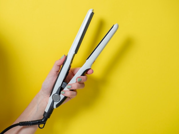 Tenaza blanca en la mano de una mujer sobre un fondo amarillo. Un accesorio para crear peinados. Belleza y moda. Cuidado del cabello.