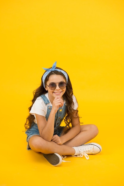 Tendencia de moda Pequeña fashionista Linda chica de moda de niño pequeño Concepto de atuendo de verano Chica de cabello largo y rizado sentarse relajante Accesorios de moda Infancia feliz sin preocupaciones Ropa moderna para adolescentes