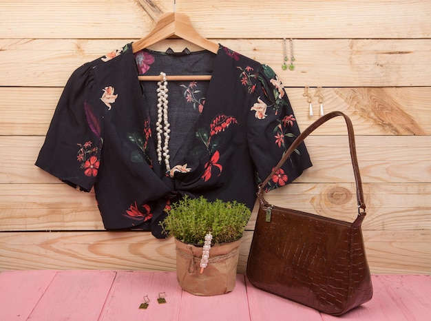 Tendências da moda - top cropped/blusa preta com estampa floral em cabide, bolsa e bijuterias: prendedor de pérolas de cabelo, colar, brincos em fundo de madeira