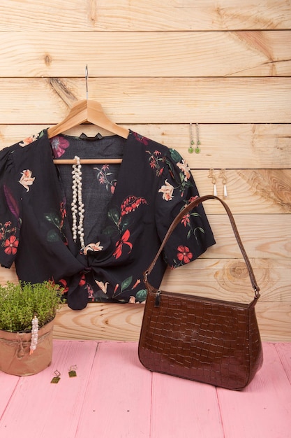 Tendências da moda - top cropped/blusa preta com estampa floral em cabide, bolsa e bijuterias: presilha de cabelo pérola, colar, brincos em fundo de madeira. foto vertical