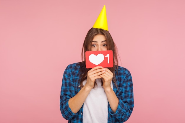 Tendencias de Internet Retrato de una chica positiva con un cono de fiesta en la cabeza sosteniendo el corazón de las redes sociales como un ícono con el blogger número uno celebrando la primera foto de estudio de suscriptor aislada en un fondo rosa