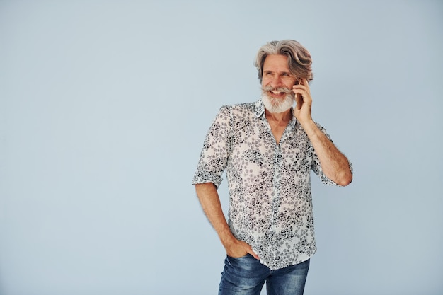 Foto tendo chamada quando em pé contra a parede homem moderno elegante sênior com cabelos grisalhos e barba dentro de casa