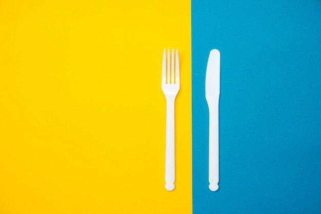 Tenedor y cuchillo de plástico blanco sobre fondo amarillo y azul. Utensilio de cocina. Vista superior. Estilo minimalista. Copiar, espacio vacío para texto