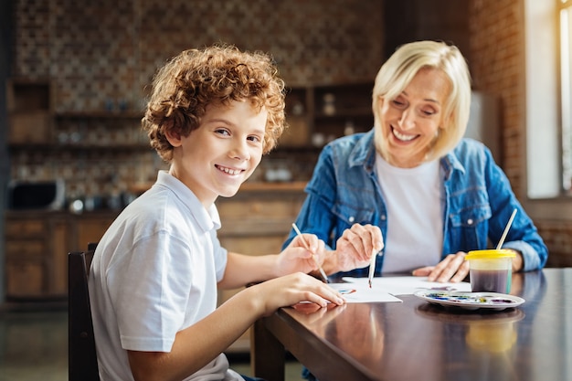 Tenemos tanta diversión. Enfoque selectivo en un nieto preadolescente inteligente que gira la cabeza hacia la cámara y sonríe mientras pasa algún tiempo con su abuela y pinta