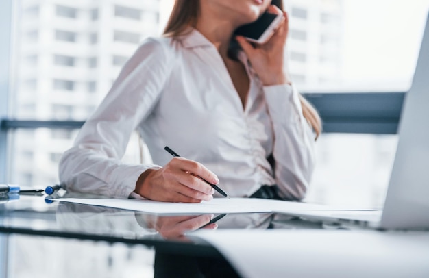 Tener una conversación por teléfono Mujer joven en ropa formal blanca está en el interior de la oficina moderna