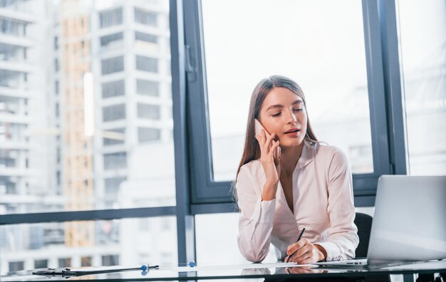 Tener una conversación por teléfono Mujer joven en ropa formal blanca está en el interior de la oficina moderna