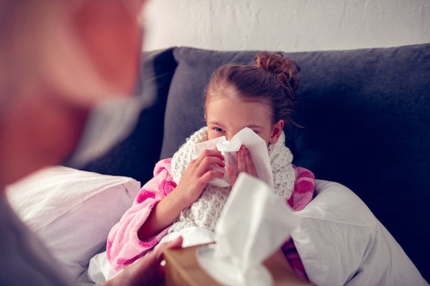 Tener moqueo. Niña de cabello oscuro que se queda en la cama con una servilleta mientras tiene goteo nasal