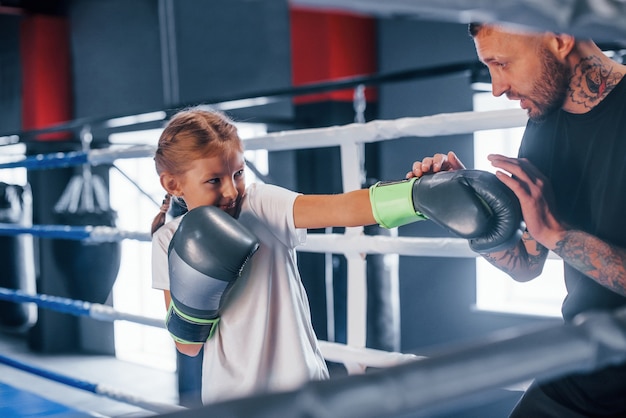 Tener sparring entre ellos en el ring de boxeo. Entrenador de boxeo tatuado joven enseña a niña linda en el gimnasio.