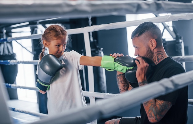 Tener sparring entre ellos en el ring de boxeo. Entrenador de boxeo tatuado joven enseña a niña linda en el gimnasio.