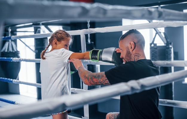 Tener sparring entre ellos en el ring de boxeo. Entrenador de boxeo tatuado joven enseña a niña linda en el gimnasio.