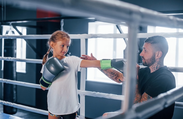 Tener sparring entre ellos en el ring de boxeo. Entrenador de boxeo tatuado joven enseña a niña linda en el gimnasio.