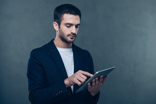 Tenho algum trabalho a fazer. Jovem bonito trabalhando em seu tablet digital em pé contra um fundo cinza