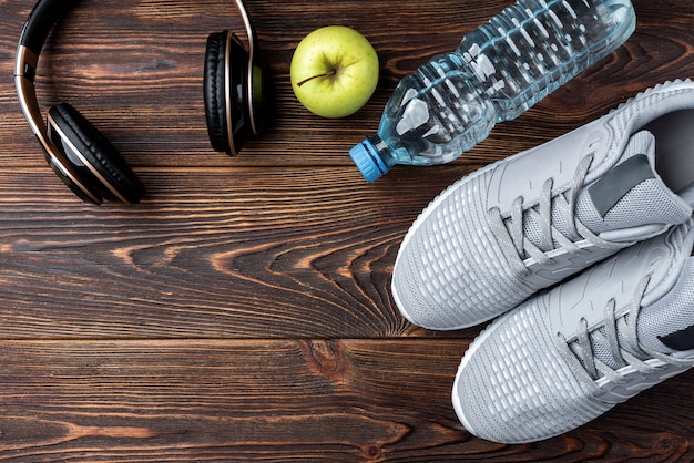 Tênis de fitness, fones de ouvido, maçã e garrafa de água no fundo escuro de madeira.