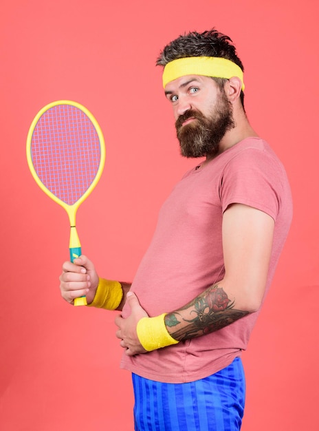 Tennis kann ein effektiver Weg sein, um Gewicht zu verlieren. Tennissport. Konzept des Tennisclubs. Der bärtige Hipster trägt ein Sportoutfit der alten Schule mit Bandagen. Athlet hält Tennisschläger in der Hand auf rotem Grund.
