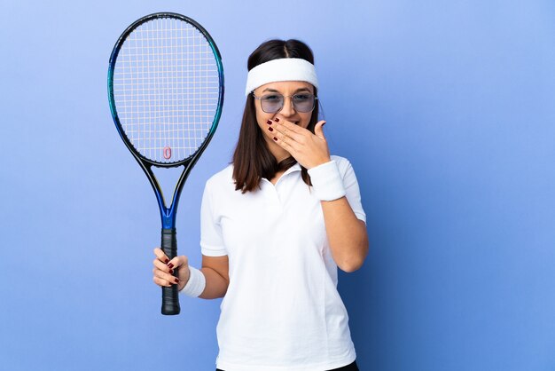 Tennisspielerin der jungen Frau über isolierter Wand glücklich und lächelnd, die Mund mit Hand bedeckt