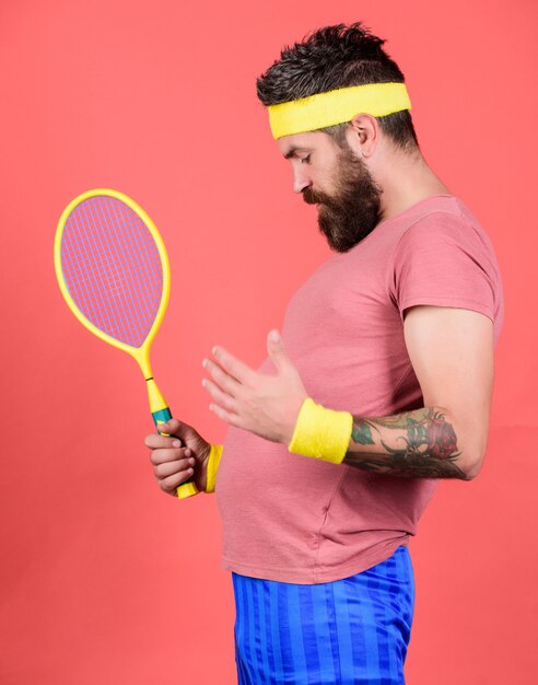 Tennissport. Der bärtige Hipster trägt ein Sportoutfit der alten Schule mit Bandagen. Athlet hält Tennisschläger in der Hand auf rotem Grund. Tennis kann ein effektiver Weg sein, um Gewicht zu verlieren. Konzept des Tennisclubs.