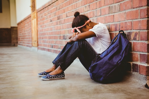 Tensed niña sentada contra la pared de ladrillo en el pasillo de la escuela