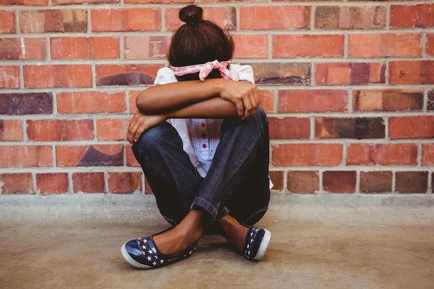 Tensed niña sentada contra la pared de ladrillo en el pasillo de la escuela