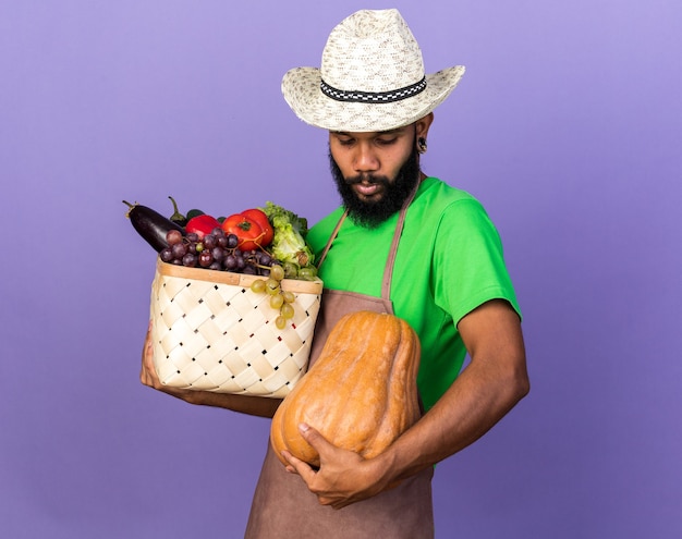 Tenso com cabeça baixa, jovem jardineiro afro-americano, usando um chapéu de jardinagem, segurando uma cesta de vegetais com abóbora isolada na parede azul