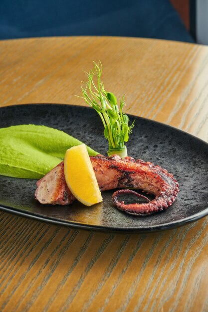 Tentáculo de pulpo a la parrilla con batido verde y salsa picante en un plato negro sobre una pared de madera. Primer plano, enfoque selectivo