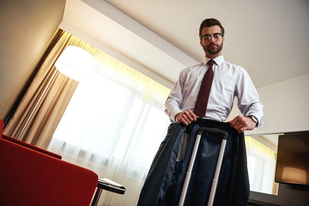 Tentando não amassar a jaqueta. Homem de negócios de óculos leva sua jaqueta para uma mala perto de um sofá vermelho em um quarto de hotel