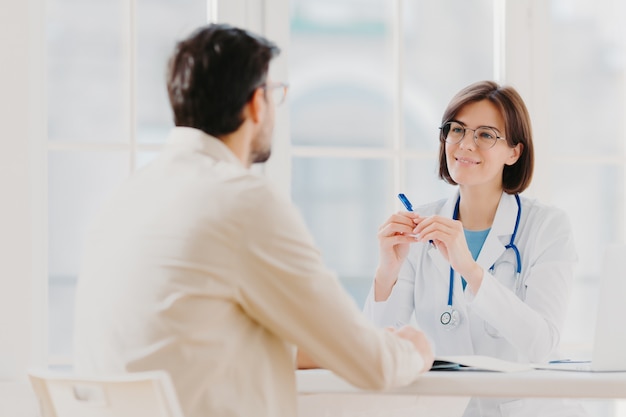 Terapeuta experto en uniforme blanco habla con el paciente, escribe en el historial médico