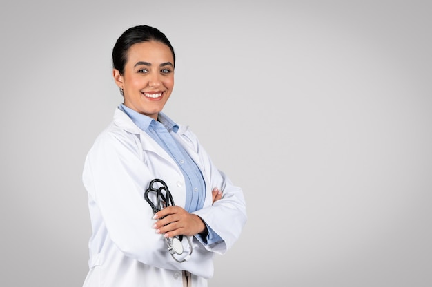 Terapeuta latina confiante sorridente segurando estetoscópio posando com braços cruzados cinza
