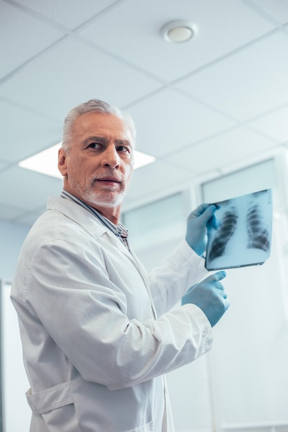 Terapeuta masculino pensativo curioso sosteniendo una imagen de rayos X de tórax y apuntando hacia ella mientras gira