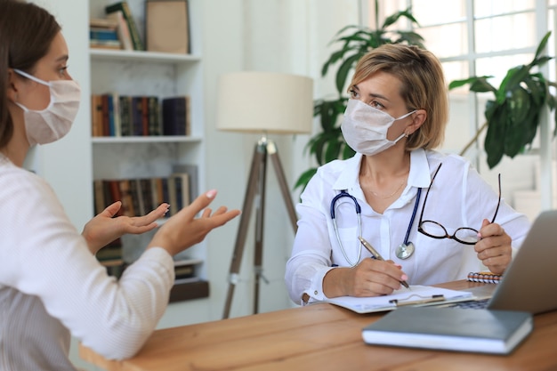 Terapeuta médica feminina de meia idade na máscara médica em consulta com o paciente no escritório.