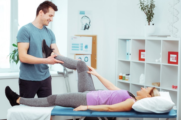 Terapeuta profissional. Jovem inteligente olhando para a paciente enquanto faz o exercício para a perna dela