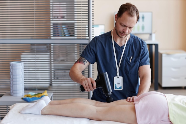 Terapeuta usando equipo médico para masajear las piernas de la paciente mientras ella está acostada en el sofá en su oficina