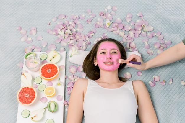 Terapia del balneario para la mujer sonriente joven que recibe la máscara facial en el salón de belleza - en el interior. Niña bonita con máscara facial rosa en la cara acostada en la cama, frutas y flores a su alrededor