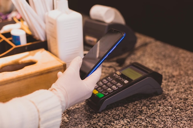 Terminal de punto de venta en el mostrador de la cafetería o restaurante Método de pago sin contacto para mantener el distanciamiento social durante la pandemia Manos de la mujer en primer plano con guantes protectores que sostienen la tarjeta bancaria cerca del terminal de punto de venta