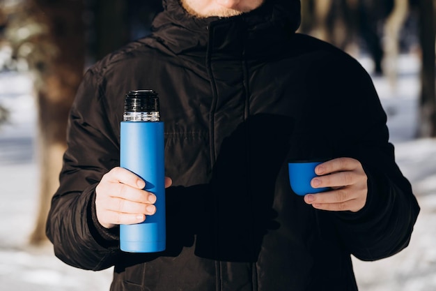 Un termo en las manos de un hombre en el bosque de invierno Copa térmica en manos masculinas