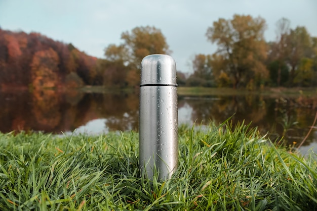 Termo de vacío de acero sobre hierba verde, bosque de otoño y lago.