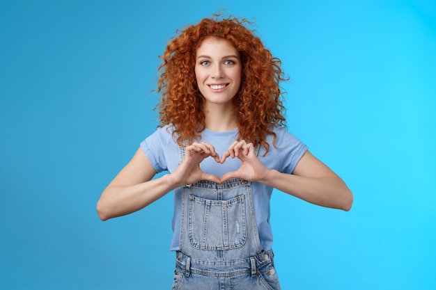 Foto ternura, amor, conceito de romance. mulher linda ruiva bonita feminina cacheada mostrar coração gesto peito expressar simpatia sentimentos apaixonados sorrindo amplamente acalentar sentimentos em pé fundo azul.