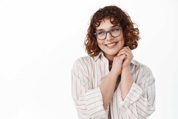 Ternura e romântica jovem sonhando acordada, imaginando algo lindo e agradável, olhando para longe e sorrindo, usando óculos, em pé no branco.