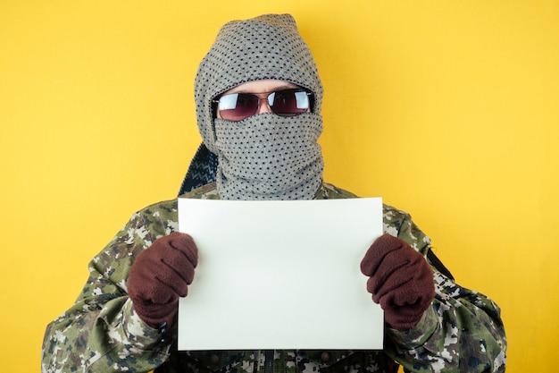 Un terrorista con un disfraz de camuflaje, gafas y una máscara sostiene una hoja de papel. el concepto de anonimato y terrorismo exige condición