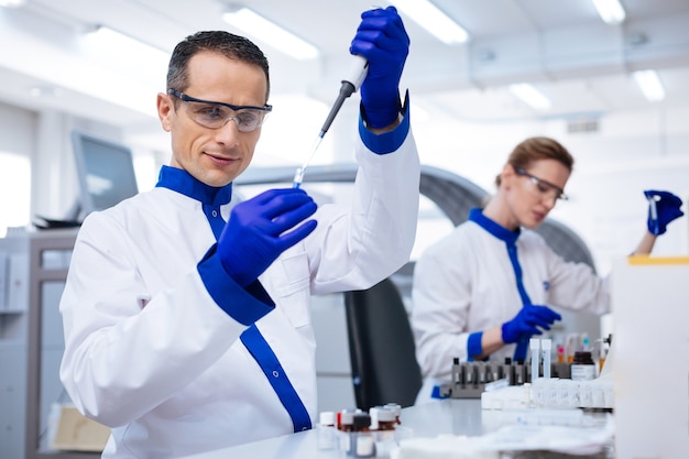 Teste de rotina. Um proeminente assistente de laboratório masculino segurando a vidraria e despejando reagente enquanto usava óculos médicos