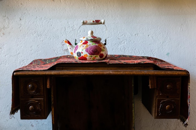 Tetera antigua decorada con flores pintadas a mano sobre una mesa antigua con un mandel bordado