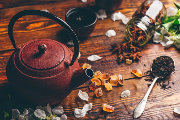 Tetera con cucharada de té, flores de manzana, azúcar y estrella de anís en mesa de madera.