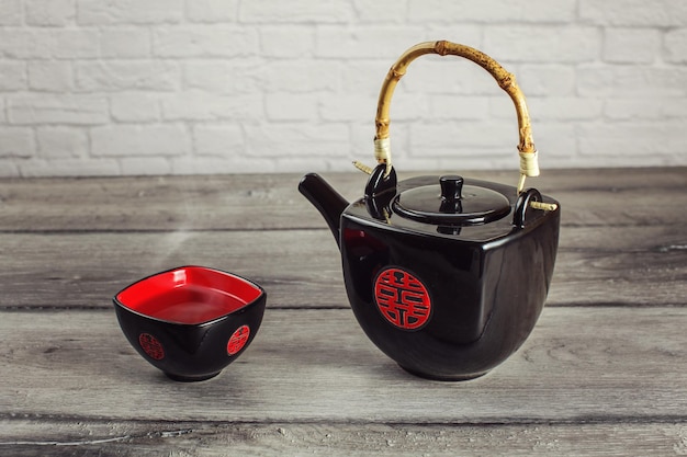 Tetera negra y taza cuadrada llena de té caliente con vapor, con un sello chino rojo que significa "Doble felicidad", colocada sobre una mesa de madera gris.