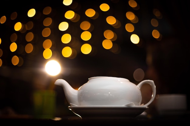 Una tetera redondeada blanca se encuentra en una barra de madera en un café nocturno. En el contexto de círculos dorados de guirnaldas amarillas de Navidad. Enfoque suave. Te de noche.